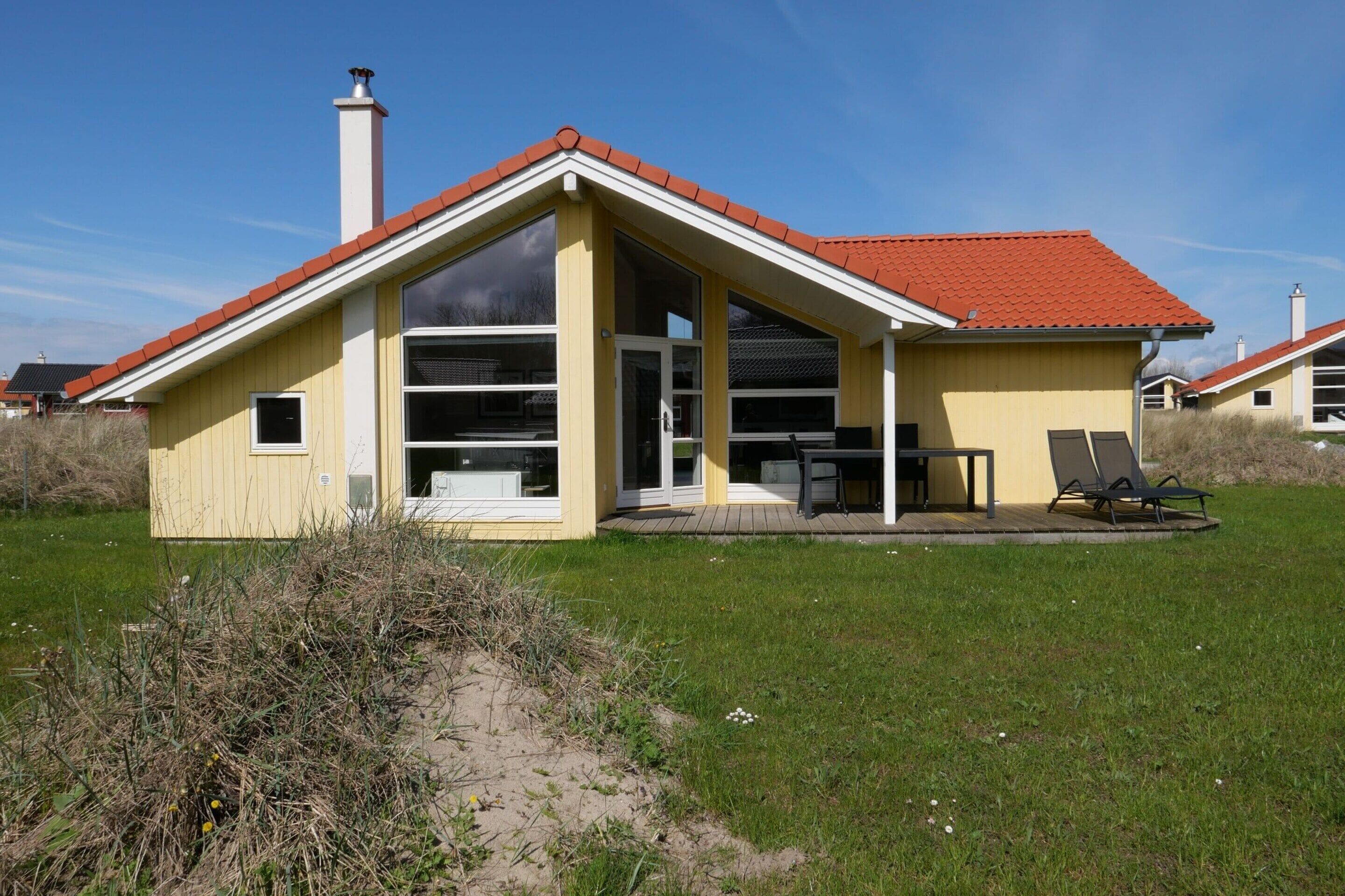 Une maison de plain-pied avec un toit recouvert de tuiles rouges, une grande fenêtre et un patio couvert aménagé avec du mobilier d’extérieur.