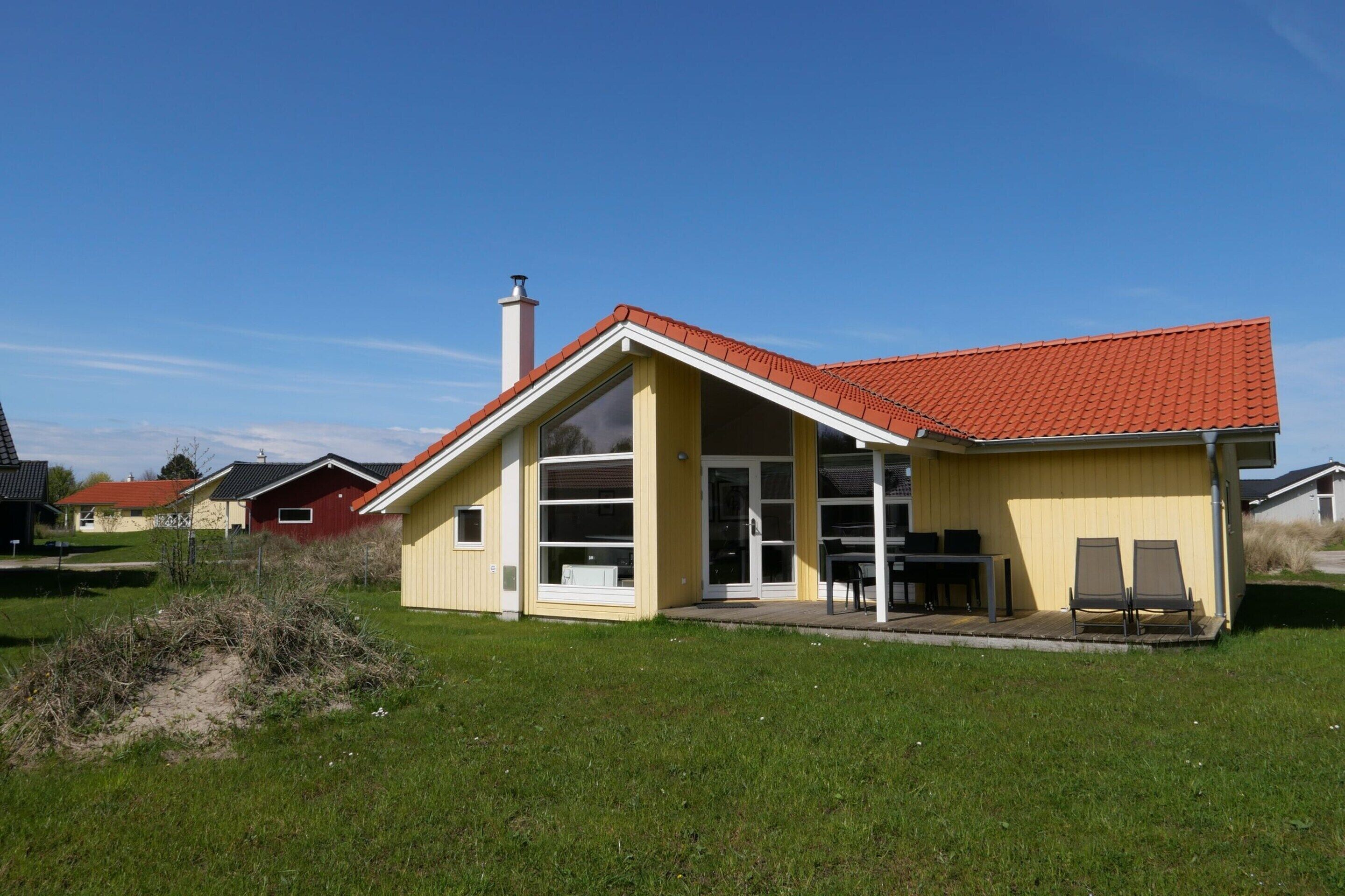Une maison de plain-pied avec un toit recouvert de tuiles rouges, une grande porte vitrée et un petit patio aménagé avec du mobilier d’extérieur.