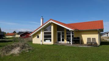 A single-story house with a red-tiled roof, a large glass door, and a small patio with outdoor furniture.