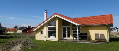 A single-story house with a red-tiled roof, a large glass door, and a small patio with outdoor furniture.