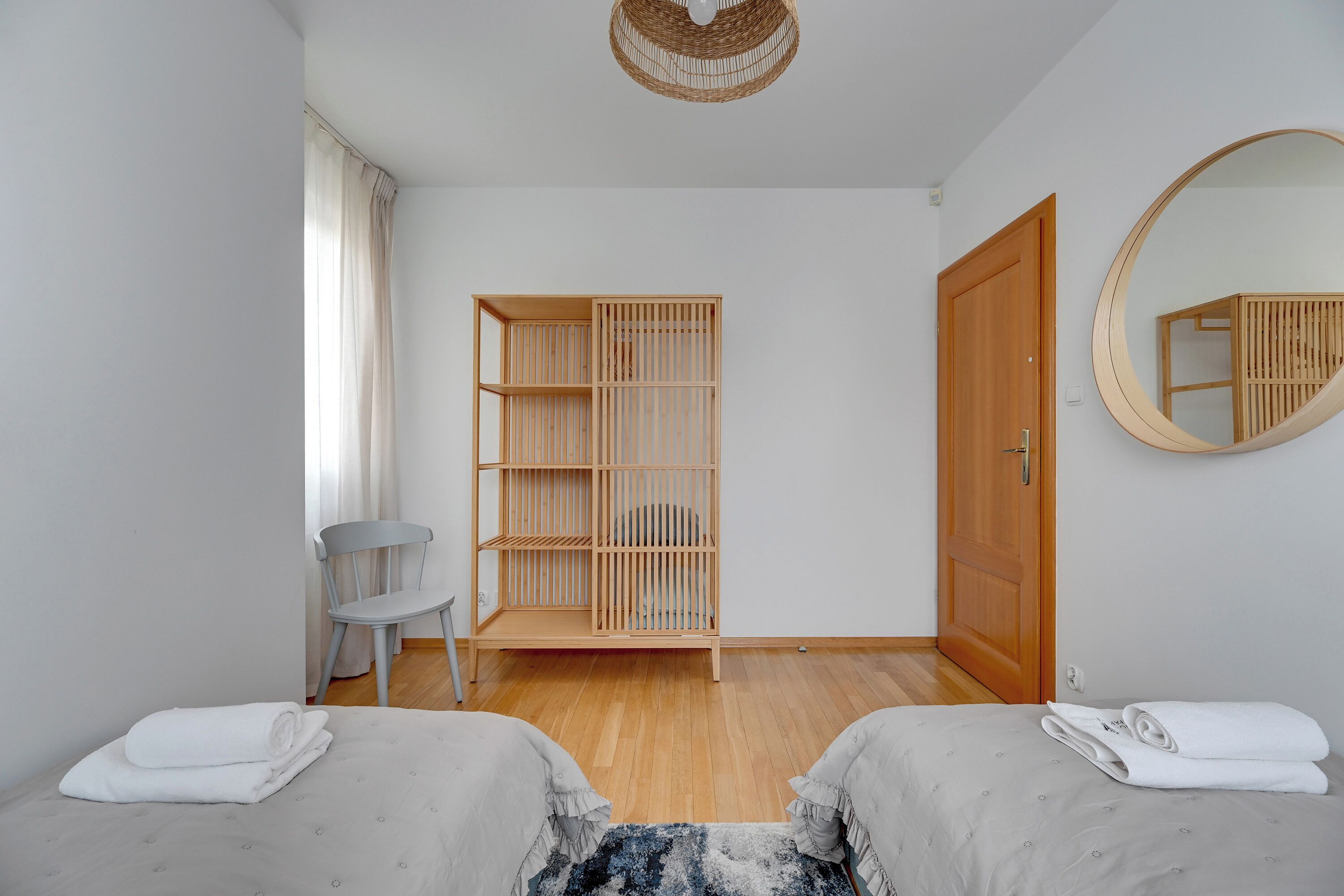 A minimalist bedroom with two beds, a wooden wardrobe, a chair, a round mirror, and a ceiling light fixture.