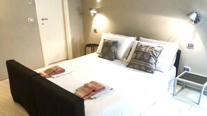 A neatly made bed with pillows and folded towels, a bedside table, and a wall-mounted light fixture. - Budello di Alassio, spacious apartment 30 meters from the beach, garage included (Alassio)
