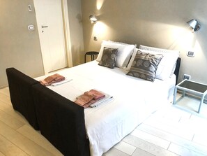 A neatly made bed with pillows and folded towels, a bedside table, and a wall-mounted light fixture. - Budello di Alassio, spacious apartment 30 meters from the beach, garage included (Alassio)