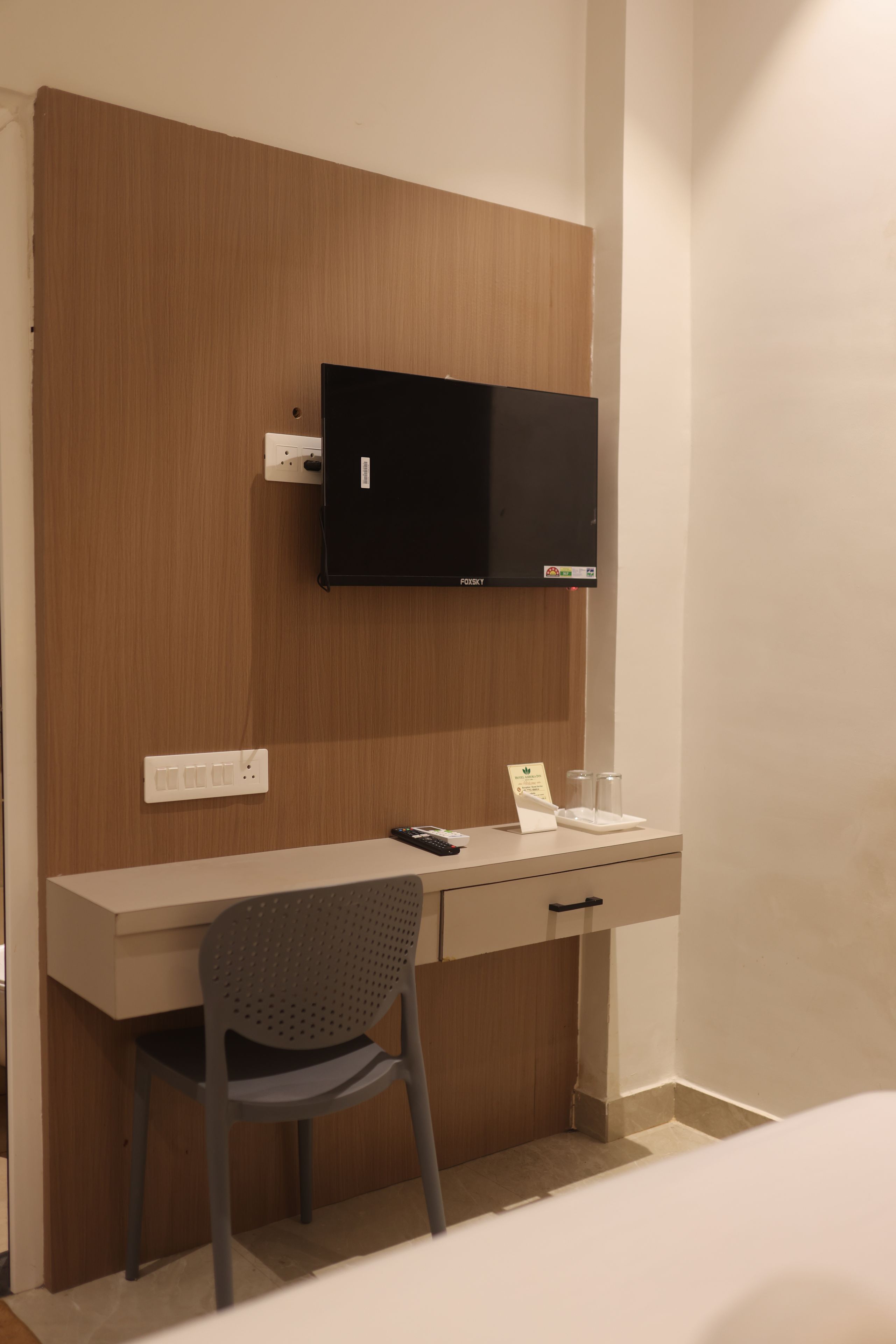 A hotel room with a wooden desk, a television mounted above it, a chair, and a small table with a remote control and a glass.