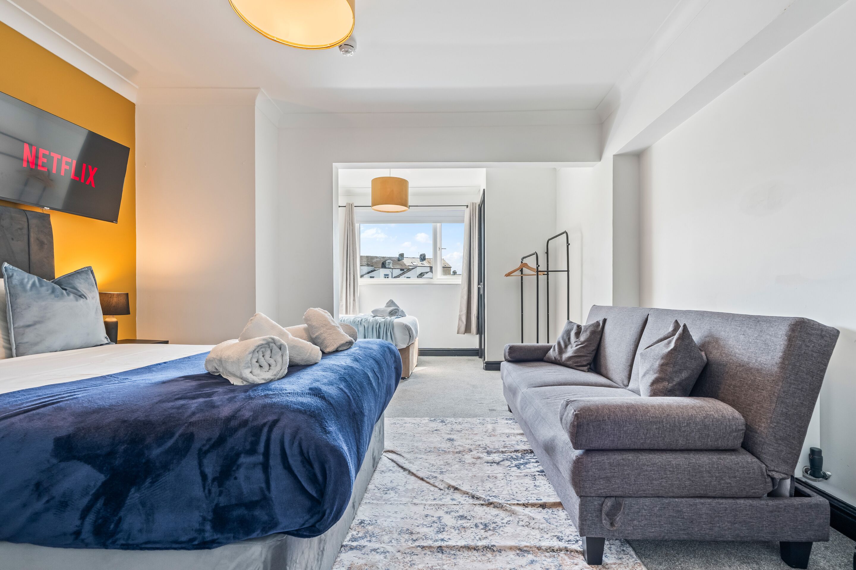A modern hotel room with a bed, a grey sofa, a TV displaying 'NETFLIX', and a view of buildings through the window.