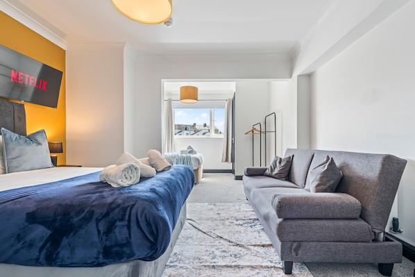 A modern hotel room with a bed, a grey sofa, a TV displaying 'NETFLIX', and a view of buildings through the window.