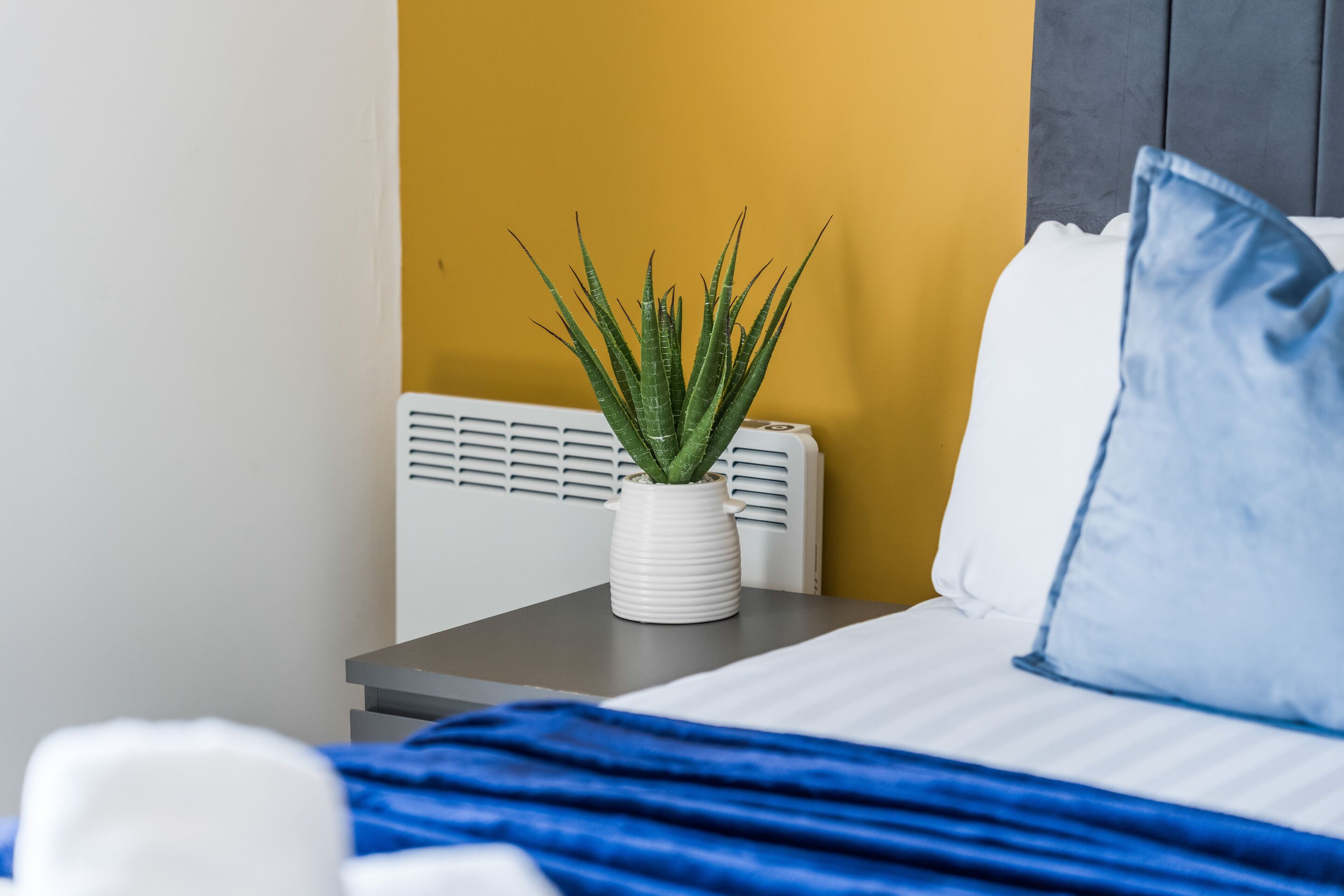 A bed with a white and blue pillow, a blue blanket, and a white bedside table with a potted plant.