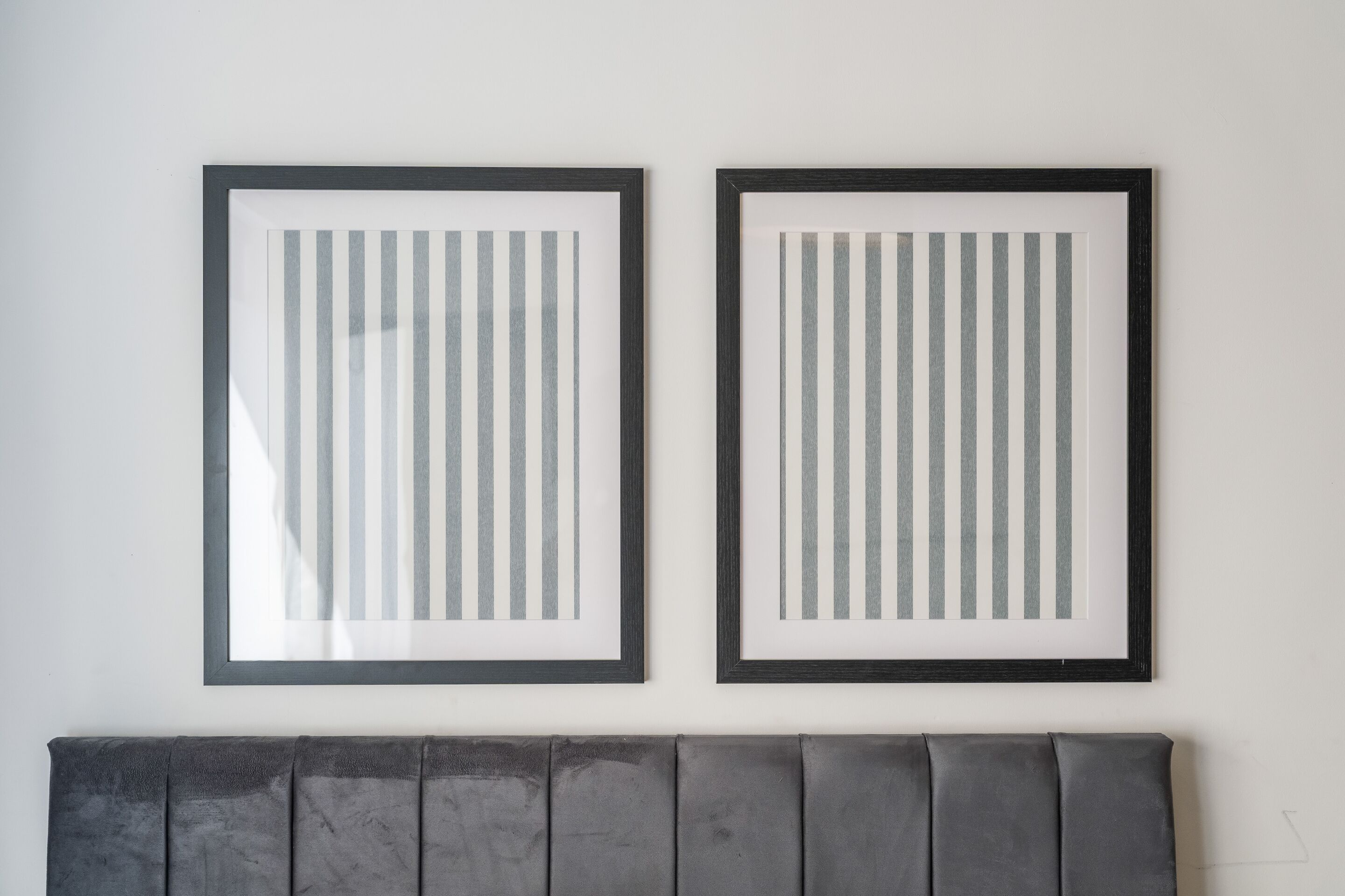 Deux œuvres d’art encadrées, ornées de rayures verticales, sont accrochées à un mur blanc, au-dessus d’une tête de lit grise recouverte de tissu.