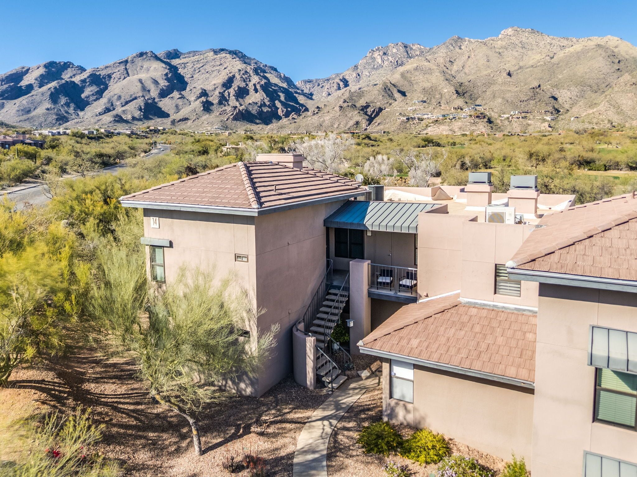 A residential area with a two-story house featuring a covered patio, a visible staircase, and a mountain backdrop.