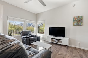 Ein Wohnzimmer mit einer Ledersofa, einem Relaxsessel, einem Couchtisch, einem Flachbildfernseher auf einem weißen Standfuß und einem Bild an der Wand.