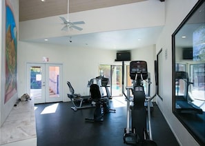 Ein Fitnessraum mit Cardiogeräten, einem Deckenventilator und einer zur Außenseite führenden Tür.