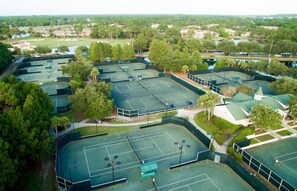 Un complesso di campi da tennis con più campi, circondato da alberi e un campo da golf sullo sfondo.