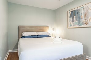 Ein ordentlich gemachtes Bett mit weißer Bettwäsche und blauen Kissen, ein gefaltetes weißes Handtuch und ein Bild an der Wand.