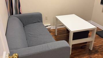 Une pièce avec un canapé gris, une table blanche, un plancher en bois, une fenêtre donnant sur des fleurs rouges et un téléviseur fixé au mur.