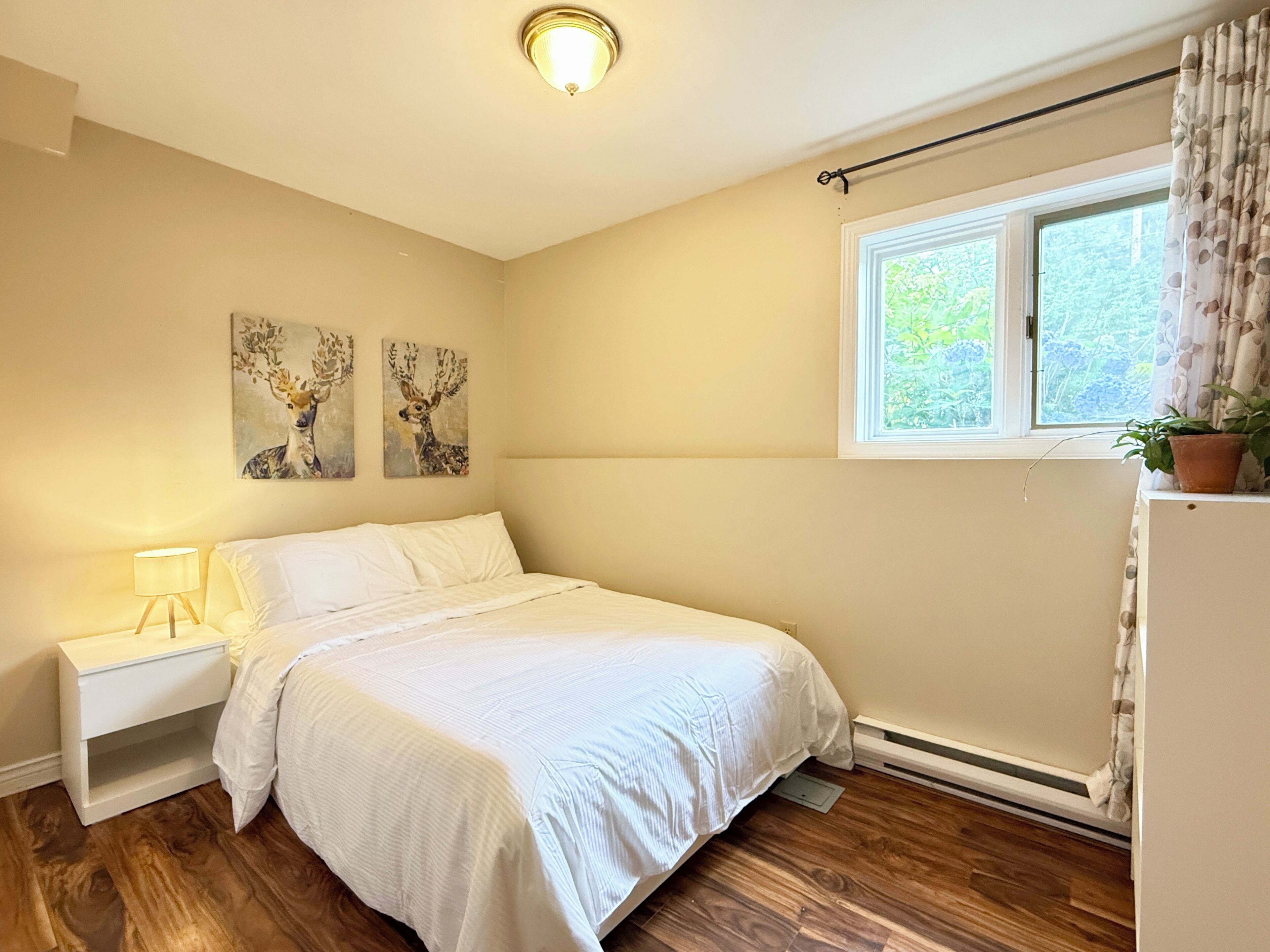 A bedroom with a bed, bedside table, lamp, wall art, window, and a potted plant.