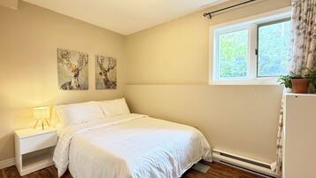 A bedroom with a bed, bedside table, lamp, wall art, window, and a potted plant.