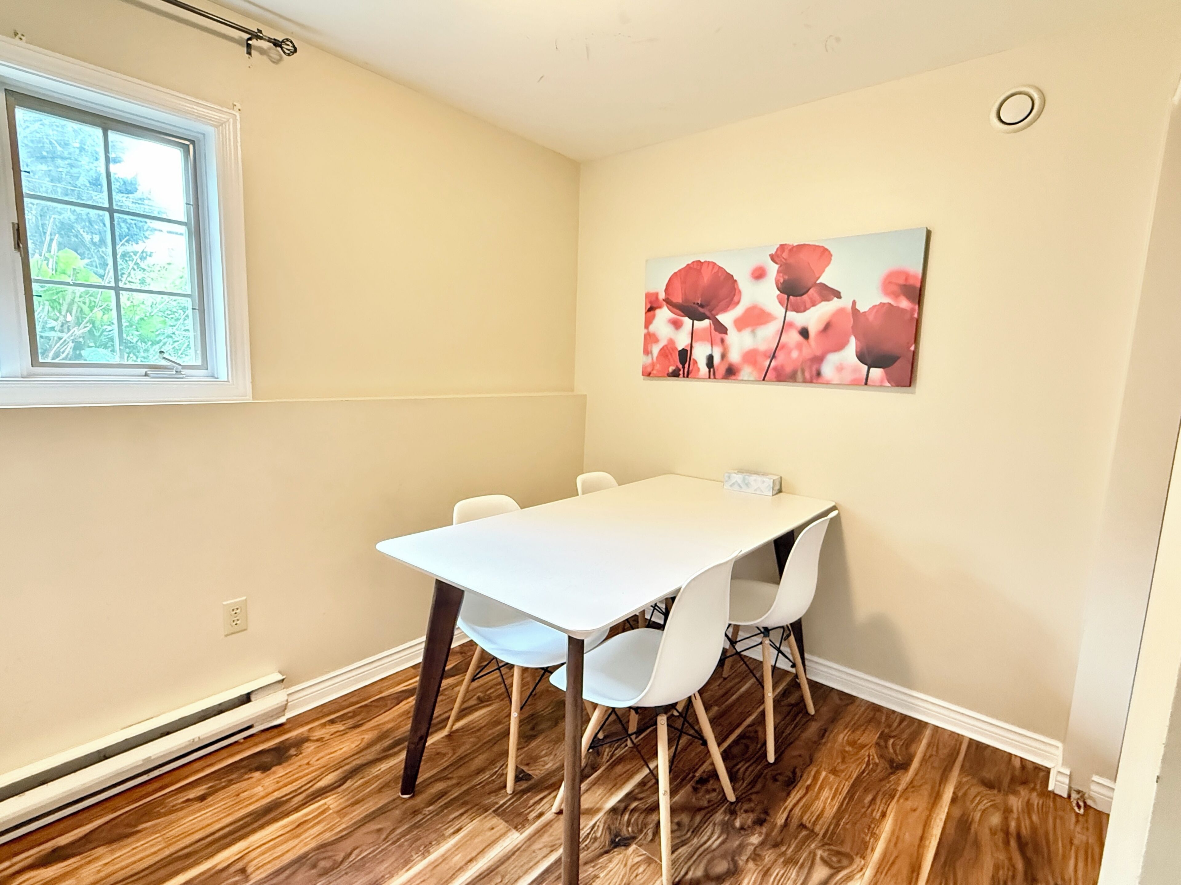 Une pièce avec une table blanche, quatre chaises, une fenêtre et un tableau représentant des fleurs rouges au mur.