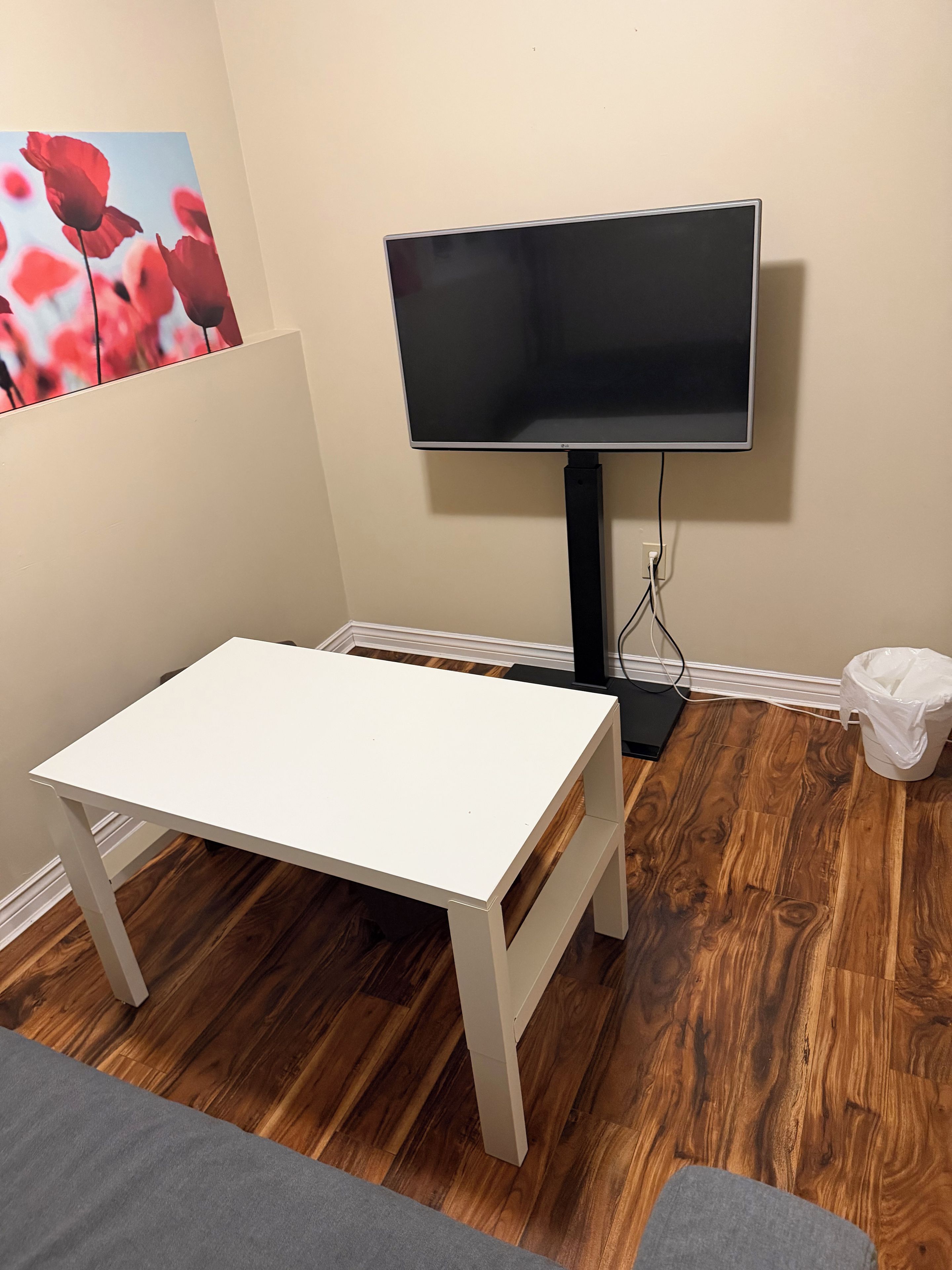 Une pièce avec une table blanche, un téléviseur posé sur un meuble, un plancher en bois et une poubelle.