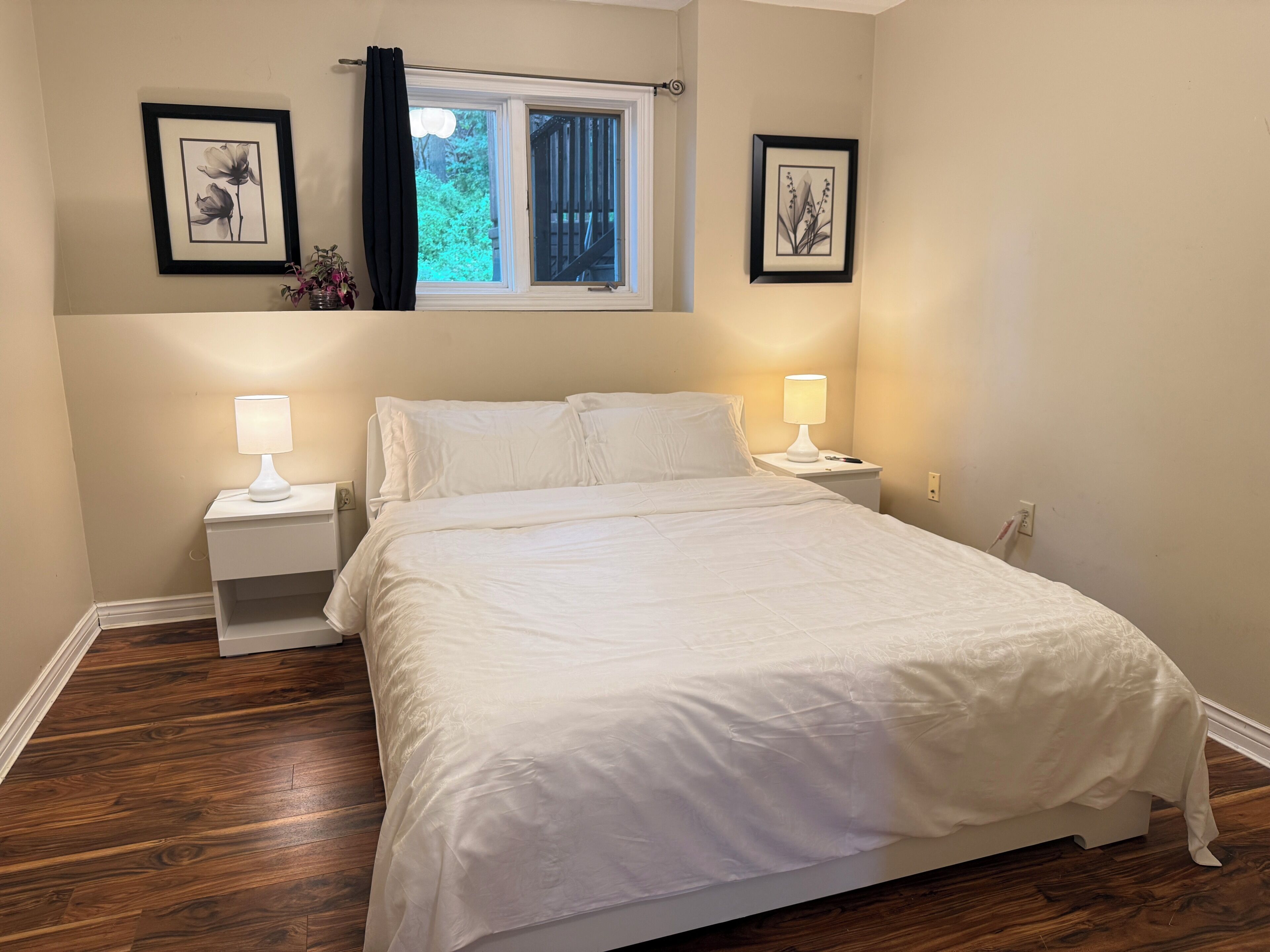 Une chambre à coucher avec un lit, deux tables de chevet avec des lampes, une fenêtre avec des rideaux et deux tableaux encadrés accrochés au mur.