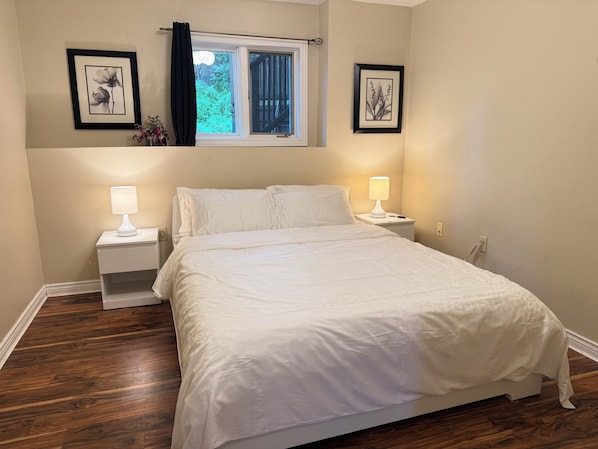 A bedroom with a bed, two bedside tables with lamps, a window with curtains, and two framed pictures on the wall.