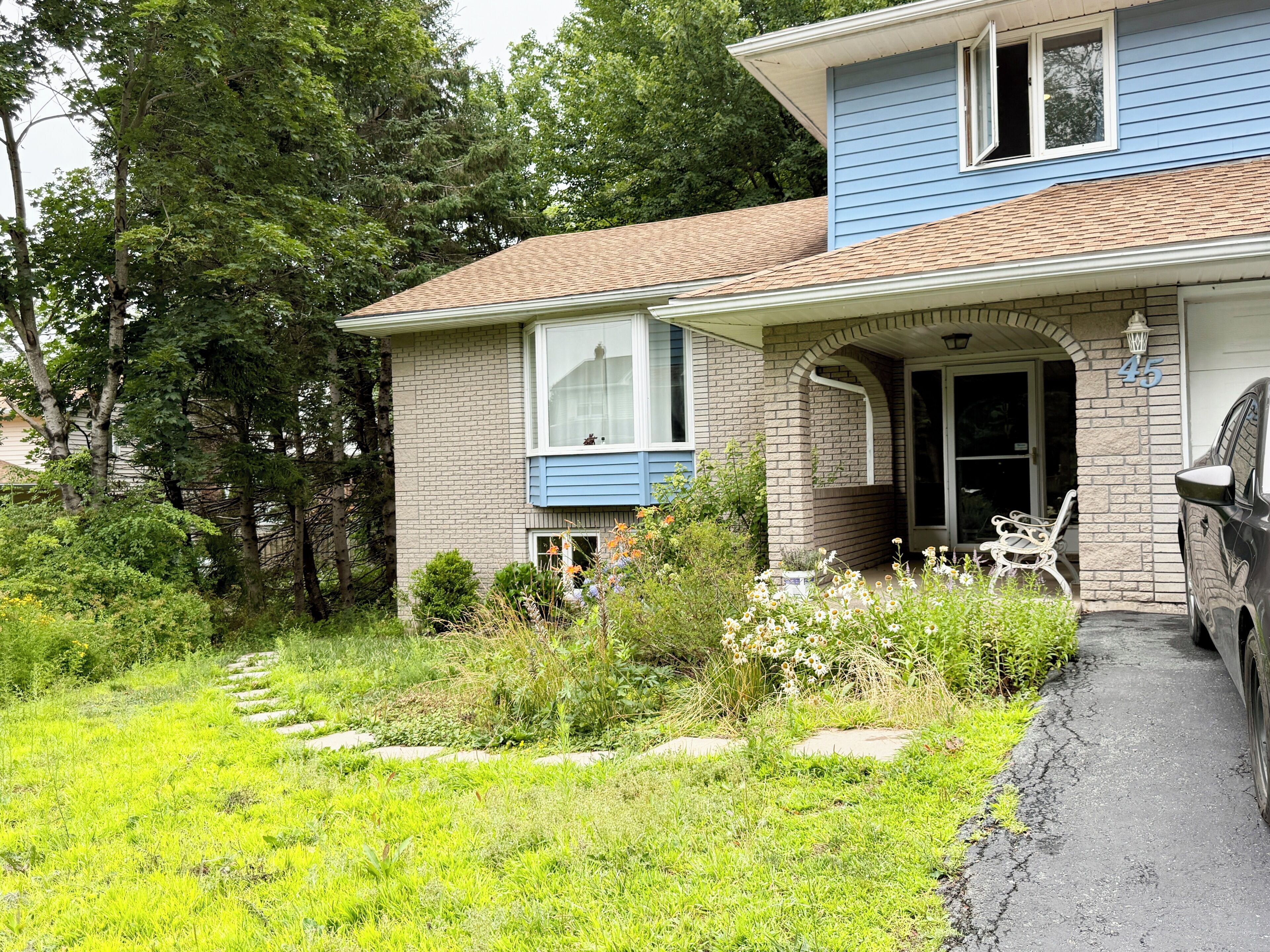 Une maison à deux étages, avec une façade en briques, un porche à l’avant et une voiture garée dans l’allée.