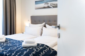 A neatly made bed with white linens and a blue blanket with cursive writing, a grey upholstered headboard, and a painting of a beach scene above the bed. - Apartment mit Dachterrasse (Lübeck)