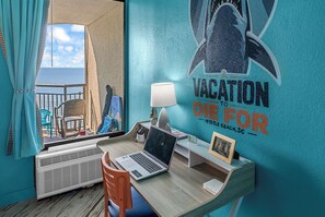 A room with a view of the sea, a desk with a laptop, a chair, a lamp, and a wall with a vacation-themed mural. - A polished oceanfront escape where subtle JAWS-inspired details, thoughtful luxury, and dog-friendly comforts create an unforgettable beach getaway. (Myrtle Beach)