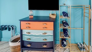 Una habitación con un televisor montado en la pared, un zapatero de madera con zapatos, una cesta de mimbre y un aparador con cajones.