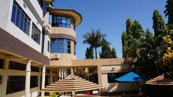 Un hôtel dont le bâtiment présente un angle incurvé, avec des espaces extérieurs aménagés avec des parasols et un jardin agrémenté de palmiers.