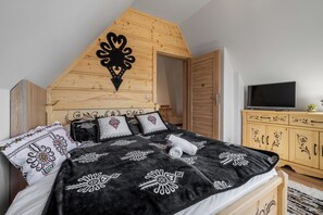 Un dormitorio con techo inclinado, una cama con ropa de cama estampada, un televisor sobre un mueble de madera y una puerta que da a otra habitación.