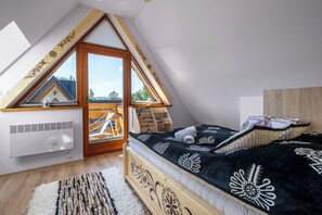 Un dormitorio con techo inclinado, una cama con colcha de estampado blanco y negro, cabecera de madera y vista de un edificio a través de una ventana.