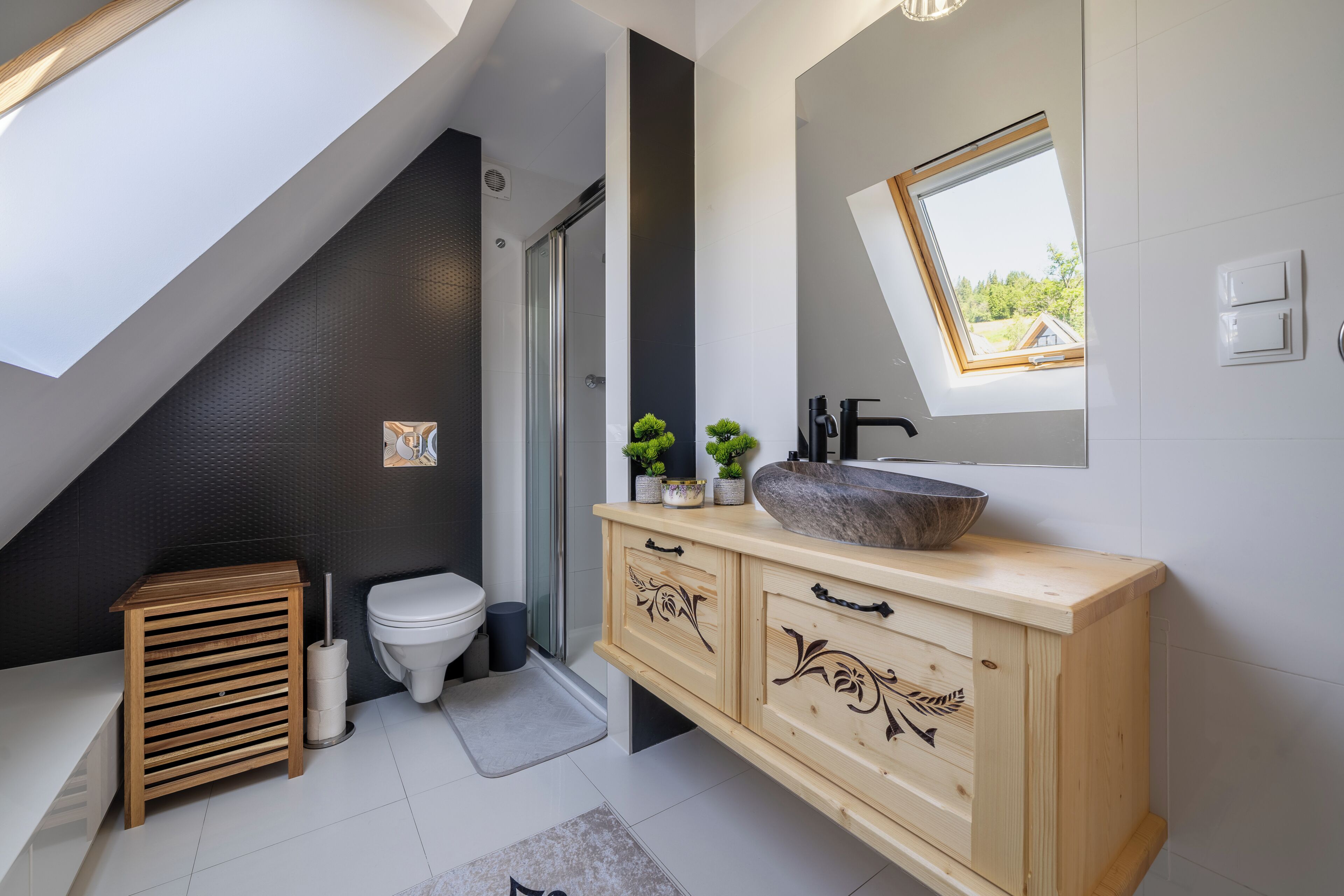 Une salle de bain moderne dotée d’une lucarne, de toilettes sur pied, d’un meuble-lavabo en bois avec un grand vasque et d’une petite plante en pot.