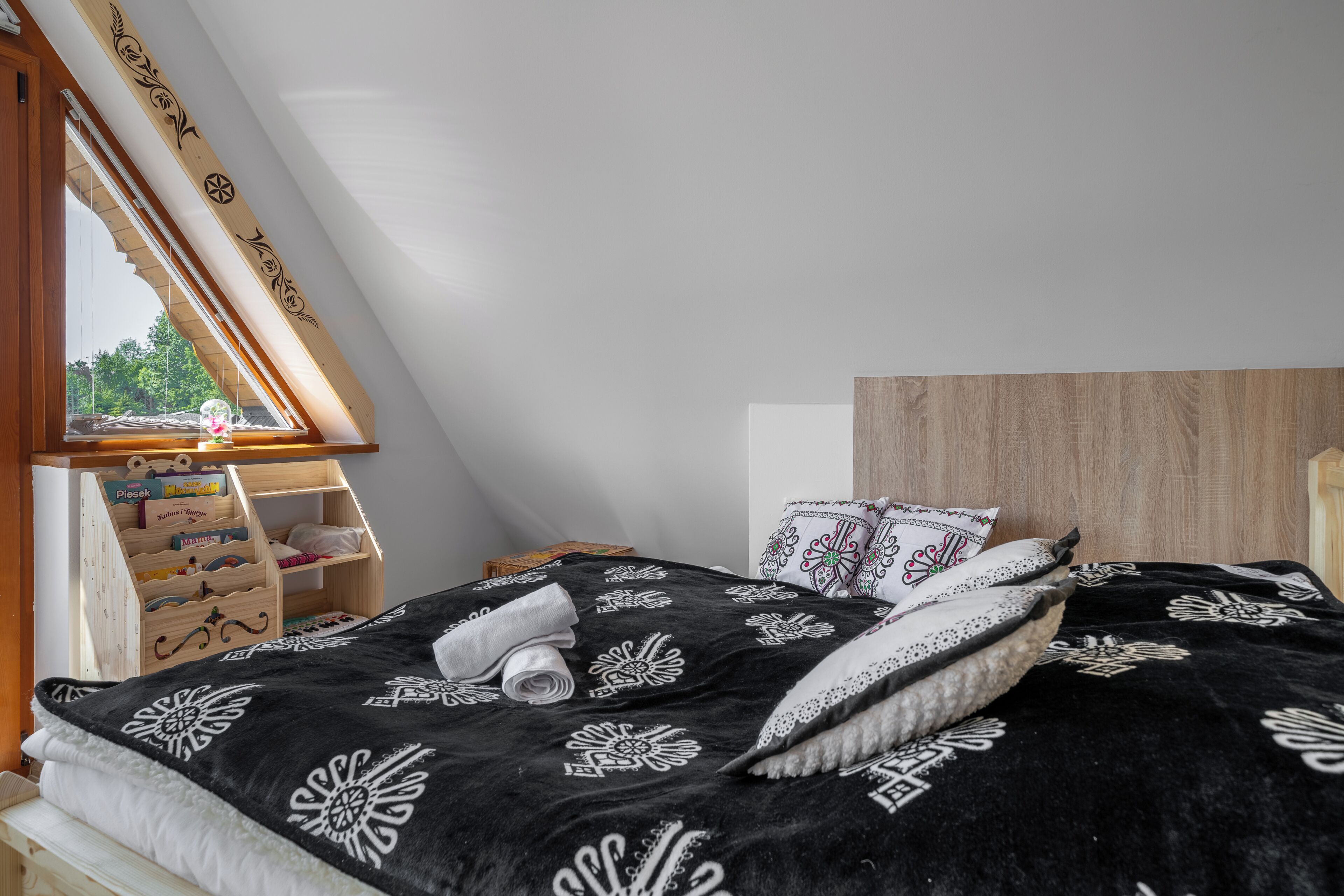 Un dormitorio con techo inclinado, cabecera de madera, cama con manta de estampado blanco y negro, y ventana con vista a follaje.