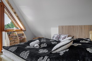 Makuuhuone, jossa on kalteva katto, puinen pääty, mustavalkoisella kuviolla varustettu peitto sängyllä ja ikkuna, josta avautuu näkymä vehreään maisemaan.