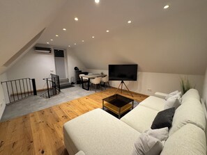 Una sala moderna con un sofá, una mesa de centro, un televisor sobre un trípode, una mesa de comedor con sillas y una escalera que sube a un nivel superior.