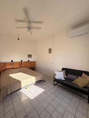 Ein Schlafzimmer mit Bett, Deckenventilator, Klimaanlage, Holz-Kopfteil, Nachttisch und einer Couch mit Kissen.