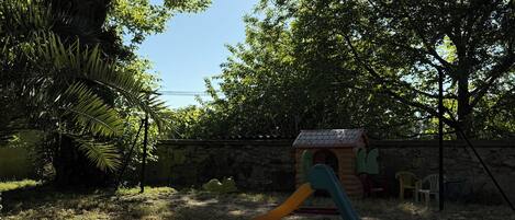A playground with a slide and a small playhouse, surrounded by trees and a grassy area.