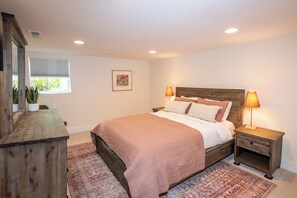 Une chambre à coucher comprenant un lit, des tables de chevet, une commode, une fenêtre avec des stores et un tableau encadré accroché au mur.