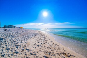Une plage parsemée d’empreintes, un ciel dégagé et des bâtiments en arrière-plan.