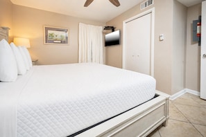 A hotel room with a large bed, a flat-screen TV mounted on the wall, a door with a fire alarm, and a framed picture on the wall. - Phoenix Iv 4042 1 Bedroom Condo (Orange Beach)