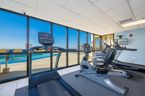 Ein Fitnessraum mit Laufbändern, Ergometer und Rudergerät, mit Blick auf einen Pool und einen Strand.