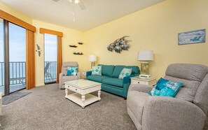 Ein Wohnzimmer mit einer Couch, Sesseln, einem Couchtisch und einer Lampe. Es gibt einen Balkon mit Vorhängen und Blickfreiheit.