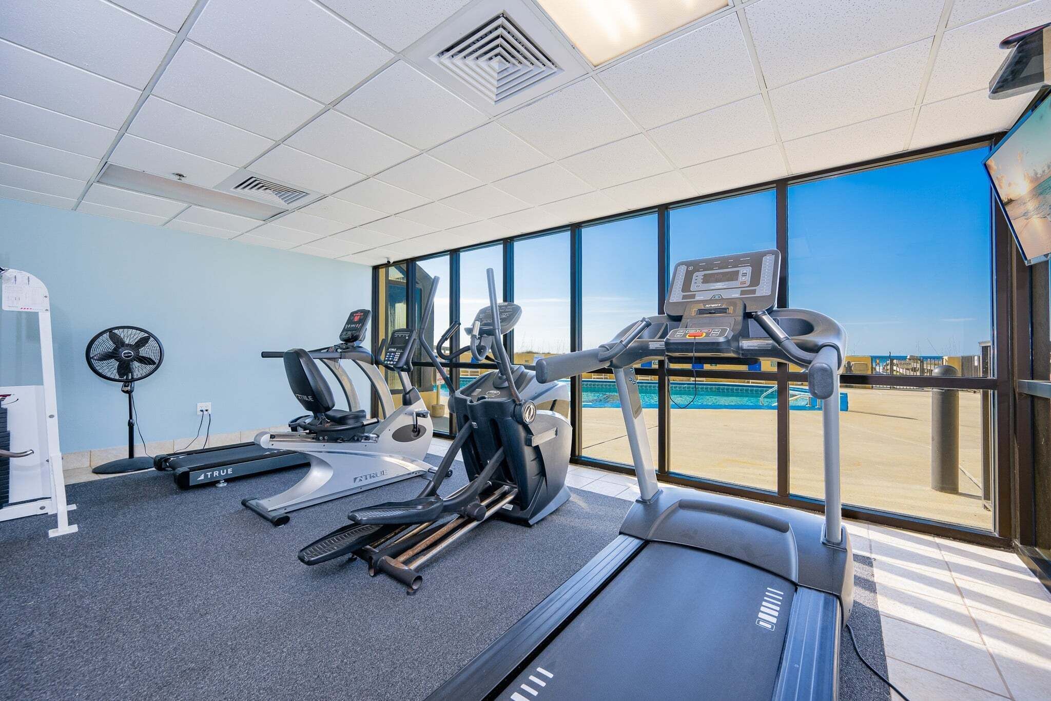 Una palestra con tapis roulant, ellittiche e una ventola, con vista su una piscina.
