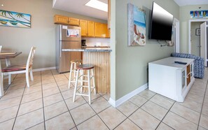 Cocina compacta con armarios de madera, refrigerador, cafetera y un pequeño comedor con mesa y sillas.