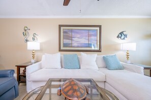 A living room with a white sofa, two side tables with lamps, a glass coffee table, and decorative fish wall art. - Phoenix Iv 4102 1 Bedroom Condo (Orange Beach)