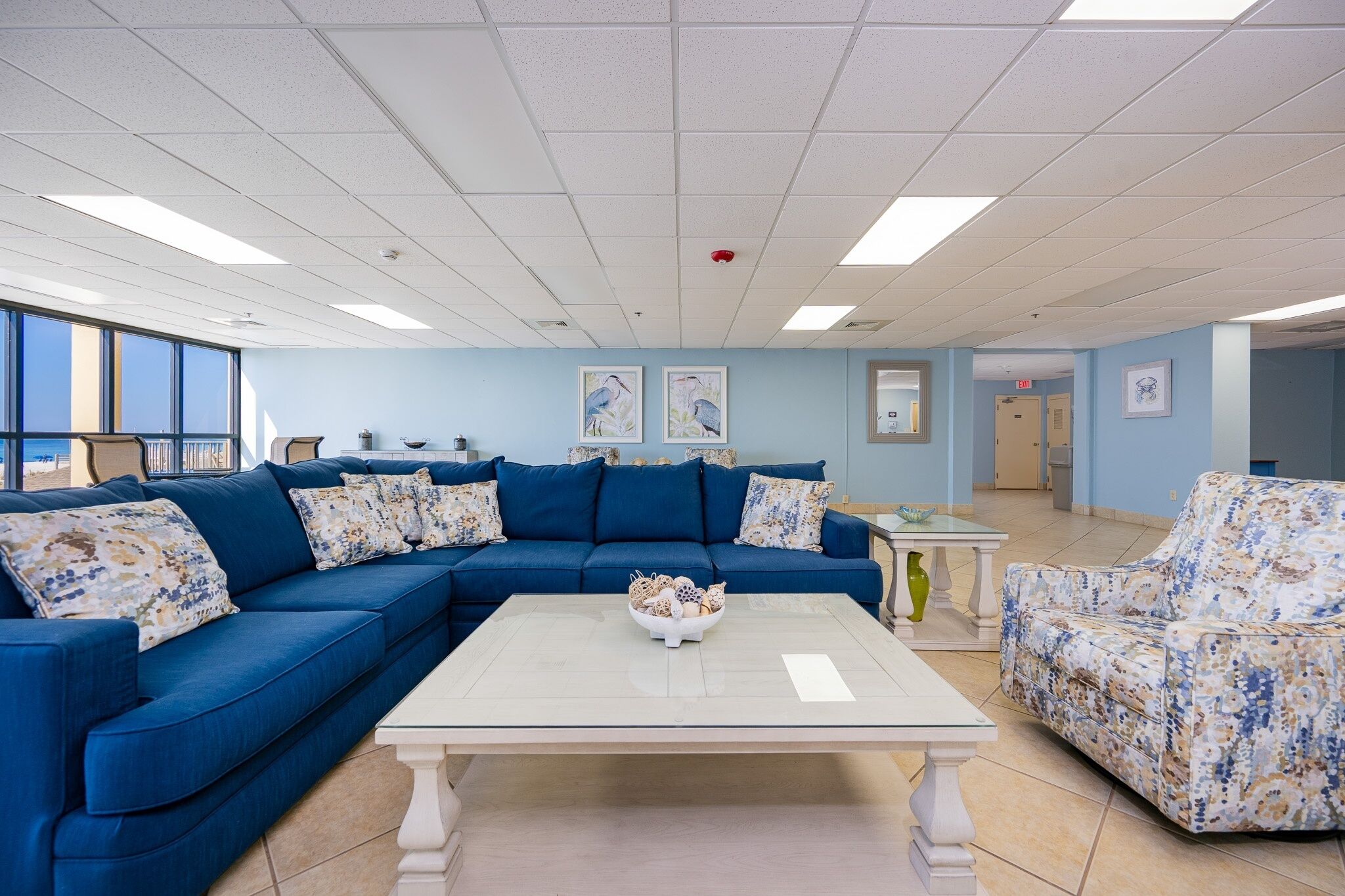 Una habitación con un sofá azul, una mesa de centro blanca y un sillón estampado. Hay cuadros en la pared y una puerta al fondo.