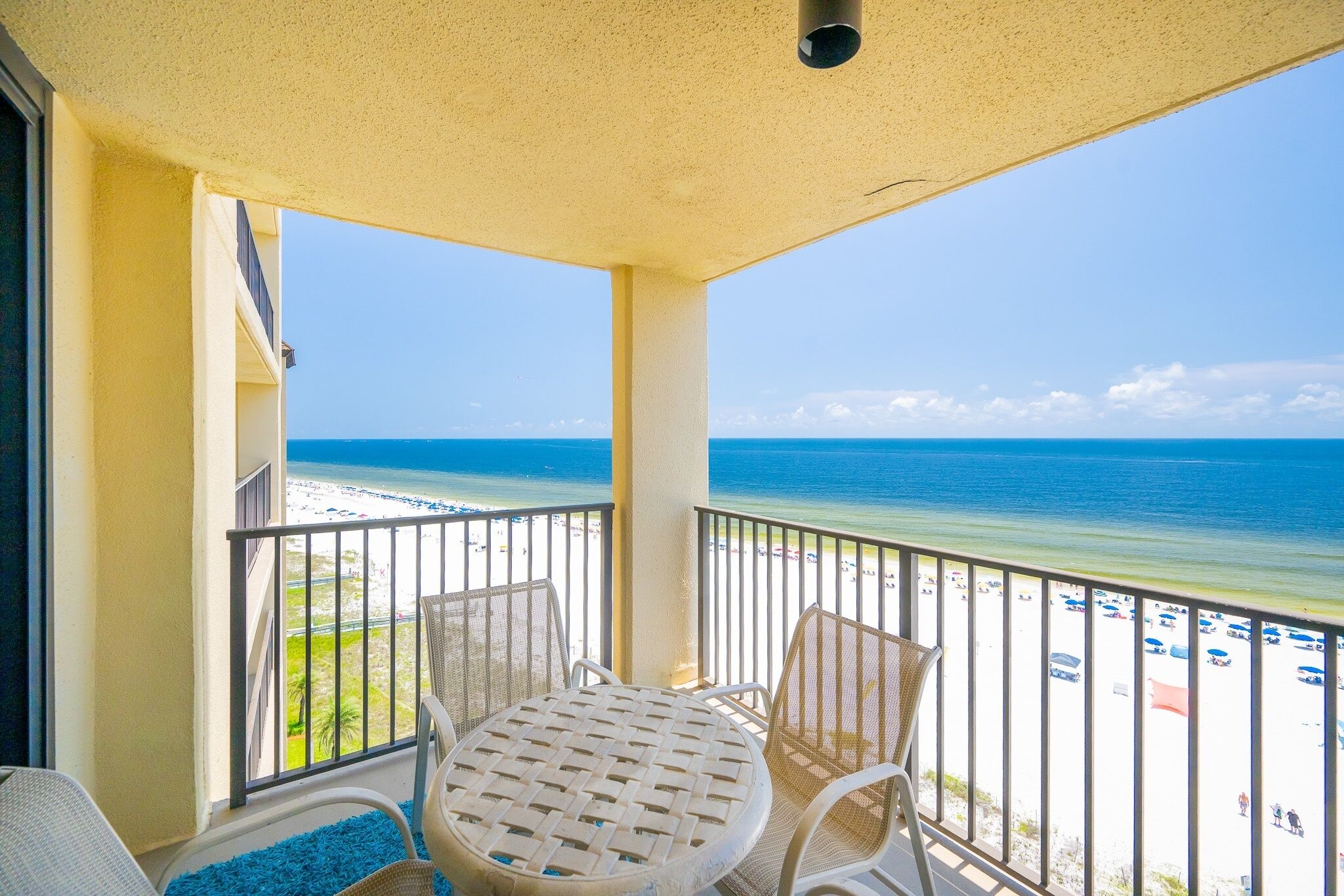 Varanda com mesa e cadeiras, vista para uma praia com guarda-sóis e pessoas.