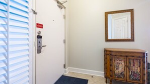 A hotel entrance with a door, a security lock, a blue mat, a wooden cabinet with a mirror, and a tiled floor. - Phoenix Ix 1202 2 Bedroom Condo (Orange Beach)