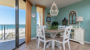A dining area with a round table and white chairs, a clock on the wall, and a view of the beach through a sliding door. - Phoenix Iv 4022 1 Bedroom Condo (Orange Beach)