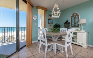 A dining area with a round table and white chairs, a clock on the wall, and a view of the beach through a sliding door. - Phoenix Iv 4022 1 Bedroom Condo (Orange Beach)